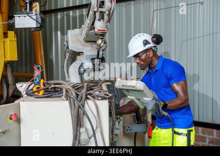 Adult African engineer uses a laptop to program an industrial robot in a workshop for automation, maintenance, and engineering concepts in Asia. Stock Photo
