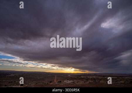 View of a vast desert landscape with a dirt road stretching toward the horizon under a dramatic dark cloudy sky at sunset Moab, Utah, United States. Stock Photo