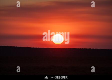 View of a bright orange sun setting over a dark desert horizon under a hazy red sky in Moab, Utah, United States. Stock Photo