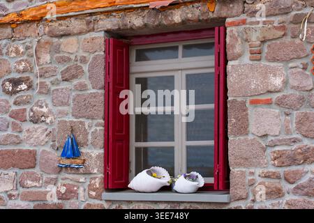 Traditional stone wall window with vibrant red shutters, seashell planters, and a small blue decorative boat ornament in the village of Skala Sikamine Stock Photo
