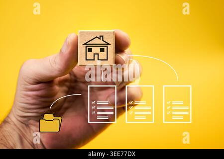 Icon of a house drawn on a wooden cube held by a hand on top of documents and a folder connected by white lines. The icons represent real estate, prop Stock Photo