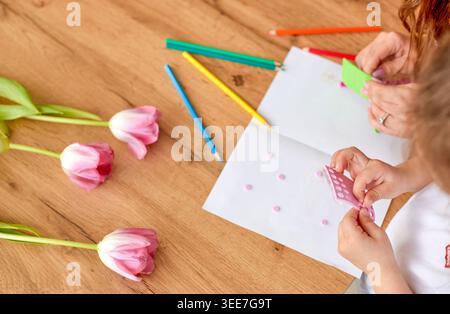 Two children create art with colored paper and markers, surrounded by pink tulips on a wooden table, showcasing a creative atmosphere Stock Photo