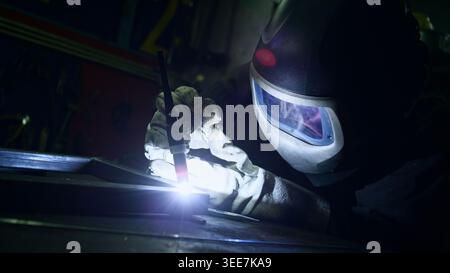 Focused and precise welder in protective workwear, face shield, and safety gloves, using a TIG welding machine in a dark workshop. Stock Photo