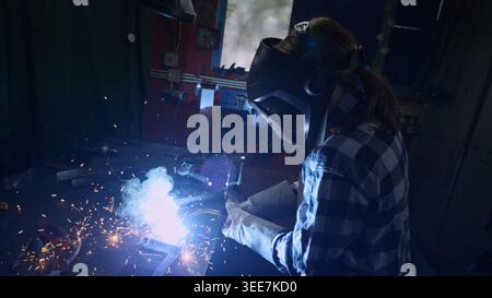 Woman welder in protective gear working with metal using a welding machine producing bright sparks, flashes, and fumes. Stock Photo