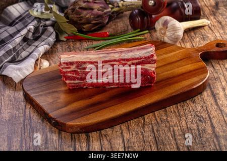 Fresh raw beef short ribs on rustic wooden cutting board surrounded by garlic, red chili peppers, green onions, bay leaves, artichoke, pepper mill Stock Photo