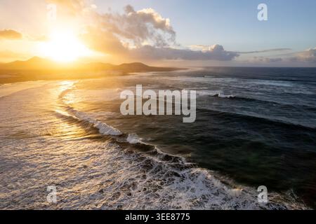 People walk along Famara beach in Lanzarote during a sunset. Waves crash on the shore while the sun sets behind distant mountains Stock Photo