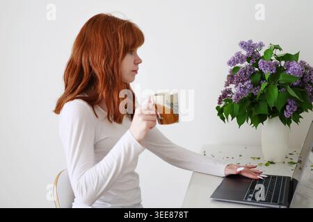 Cozy indoor spring moment with young woman at laptop smelling fresh lilac flowers, soft light and peaceful mood. Stock Photo