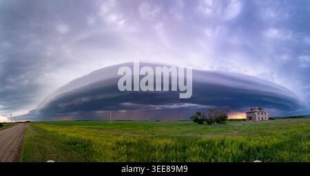 A vast, imposing shelf cloud stretches across the horizon, casting a dramatic shadow over a green field and an abandoned stone structure under a turbu Stock Photo