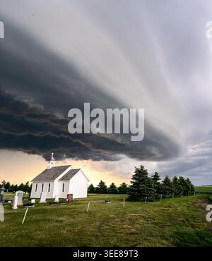 A serene white chapel stands in a graveyard beneath a vast, ominous shelf cloud formation during twilight Stock Photo