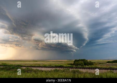 A powerful storm system with a rotating wall cloud and rain shafts approaches across a flat, grassy terrain with railroad tracks in the foreground Stock Photo