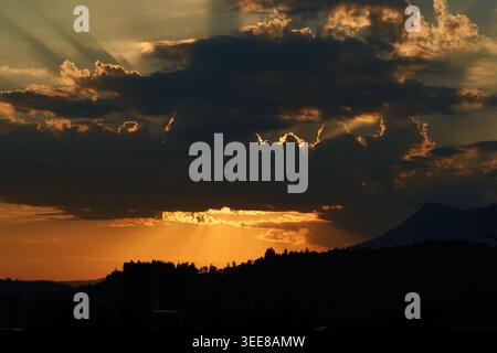 Golden sunset sunrays burst through heavy clouds, casting dramatic light over silhouetted hills and a distant mountain. Stock Photo