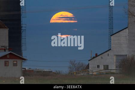 The full moon glows orange as it rises behind silhouetted farm structures and utility towers against a twilight sky Stock Photo