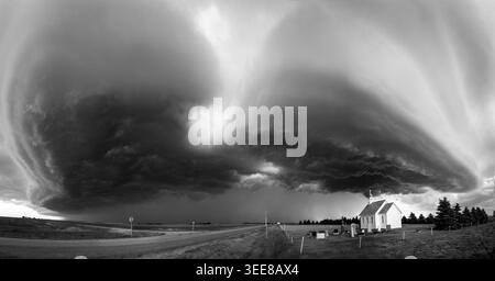 A panoramic black and white view captures a supercell thunderstorm forming over a rural landscape with a small church and graveyard Stock Photo