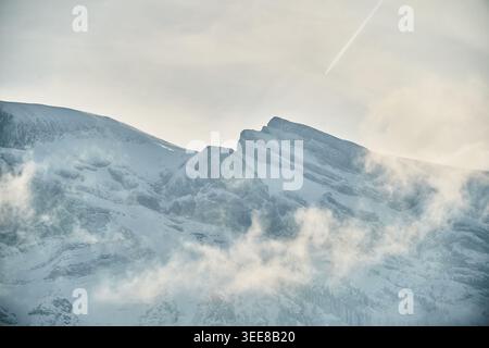 Snow-covered alpine ridge emerges through low clouds and mist under a pale sky with a contrail. Stock Photo