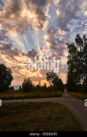 Dramatic sunset sky with sun rays breaking through clouds above a quiet country road and trees. Stock Photo