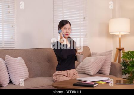 Asian woman talking on phone while sitting on sofa at home living room working remotely during business call Stock Photo