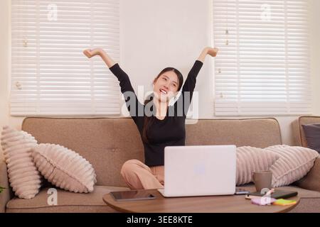 Happy Asian female celebrating achievement success working from home with laptop stretching arms on couch feeling Stock Photo