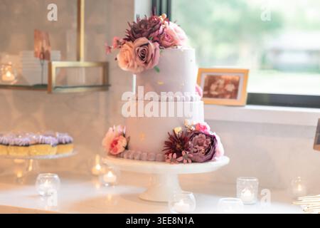 Two-tier white celebration cake decorated with pink and burgundy roses and dahlias on a cake stand, with frosted cupcakes and candlelit votives Stock Photo