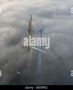 Aerial view of a wind turbine under construction with a tall yellow crane rising above a thick layer of white fog in Netherlands. Stock Photo