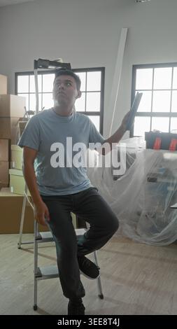 Man holding tablet while seated on a step ladder amid moving boxes, toolbox and plastic covered furniture in building; reflective. Stock Photo
