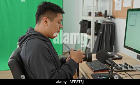Man sits at desk adjusting camera lens in studio with green screen, keyboard, microphone and monitor visible; quiet calm. Stock Photo