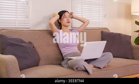 Young Asian women relaxing stretching arms with laptop on sofa taking break from remote work at cozy home living room Stock Photo
