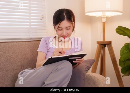 young Asian women writing in notebook while sitting comfortably on sofa at home living room planning organizing daily Stock Photo