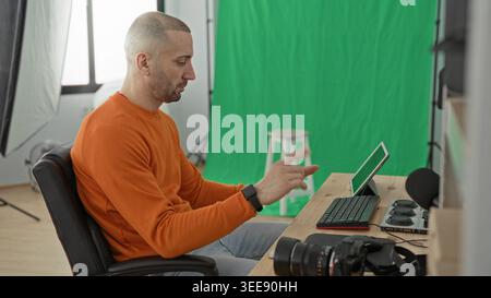 Man photographer pointing at tablet and gesturing with hands at desk in studio, green screen and camera visible; concentration creativity. Stock Photo