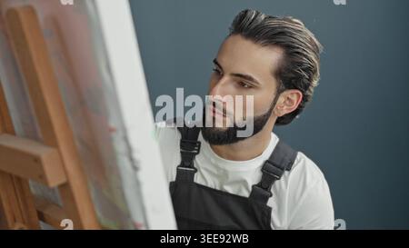 Man painter in apron looks intently at canvas on an easel in studio, head tilted studying paint marks while wearing white shirt and black apron; conce Stock Photo