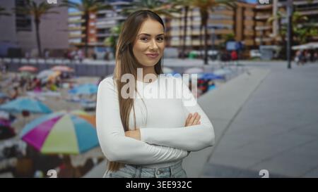 Woman with arms crossed and slight smile wearing a white long sleeve top on a street promenade by beach umbrellas and palm trees; confidence empowerme Stock Photo