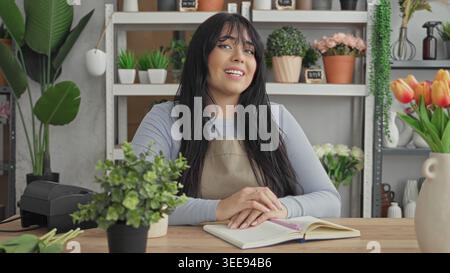 Woman florist in apron smiling with hands clasped on open notebook at studio counter, surrounded by potted plants, vases, shelves and tulips; welcomin Stock Photo