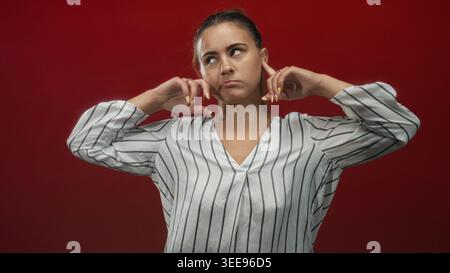 Woman plugging ears with fingers in studio against a deep red backdrop, wearing a white striped blouse and looking upward with hands raised; frustrati Stock Photo