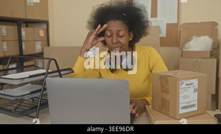 Woman with laptop, stacked parcels and headphones, hand on temple while sorting boxes at a packing building; stress overwhelm. Stock Photo