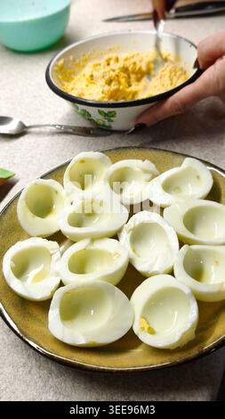 Hard-boiled egg halves prepared for filled with creamy mixture on plate, hand mixing filling in bowl, eggs and preparation visible in kitchen, closeup Stock Photo