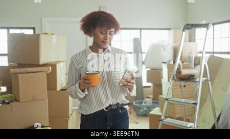 Woman holds smartphone and orange mug, hand tapping phone while surrounded by moving boxes in building; settling in contentment. Stock Photo