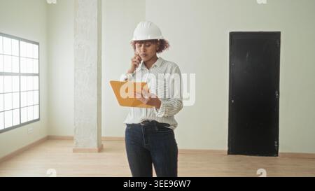 Woman latin architect with hardhat and clipboard holding smartphone to ear while reading notes in an empty building under inspection; focused inspecti Stock Photo