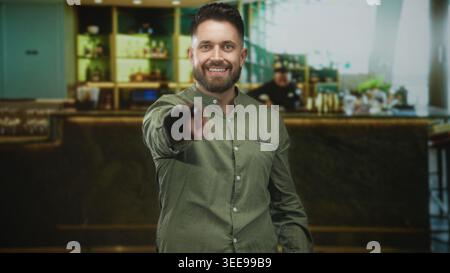 Young hispanic man with beard points finger at camera inside a bar building, smiling and leaning forward; confidence welcome. Stock Photo