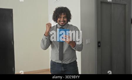 Man holding blue tablet, fist pump while smiling in building hallway wearing shirt and tie checking property listing on screen; joyful success. Stock Photo