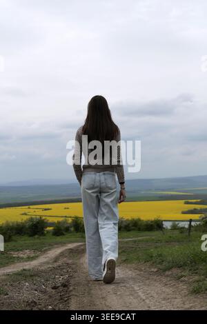 Young woman walking away on dirt road, observing yellow canola field under cloudy sky. Back view, contemplating nature outdoors Stock Photo