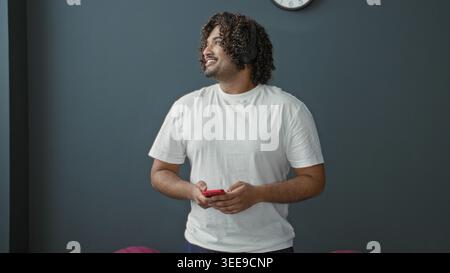 Man tapping red smartphone with fingers while standing in studio gym, wearing white tee and focused on screen; calm attentive focus. Stock Photo