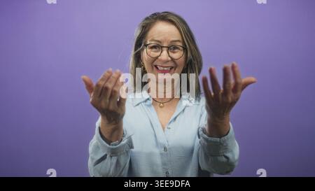 Hispanic woman with grey hair beckons with both hands toward camera in a studio wearing glasses and a light blue shirt; friendly invitation. Stock Photo