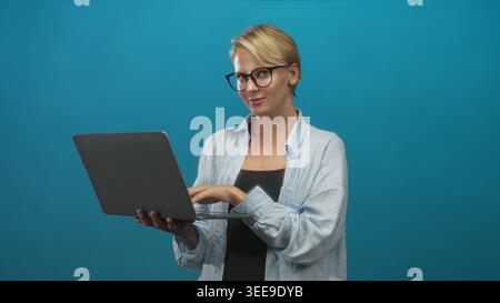 Blond caucasian woman typing on laptop in studio, holding the device with both hands and looking at the screen while wearing glasses and a light shirt Stock Photo