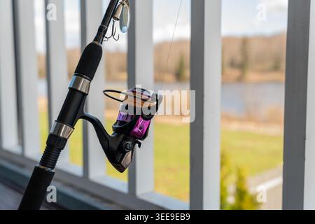 Spinning Rod Hanging By Window Overlooking Blurred Lake And Reeds, Purple And Gold Reel Perched On Railing, Ready For Quick Cast, Lifestyle Scene Stock Photo
