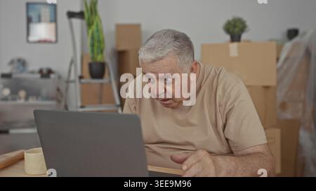 Man holding head with hands over laptop among packed moving boxes in building; moving paperwork frustration. Stock Photo