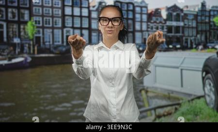 Woman in white shirt and glasses beckons with hands by a canal on a street; urban invitation interaction. Stock Photo