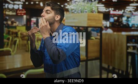 Man worker in blue work overalls and uniform with beard blows a kiss with hand to mouth near wooden tables and chairs in a cafe building; affection go Stock Photo