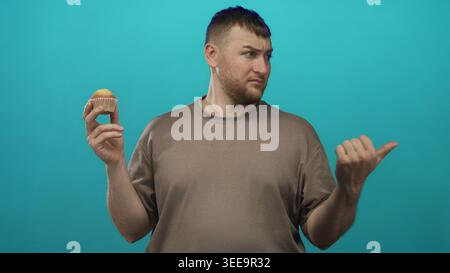 Man holding muffin with thumb pointing sideways in studio with teal backdrop, young heavyset wearing brown tee and puzzled frown; doubt hesitation. Stock Photo