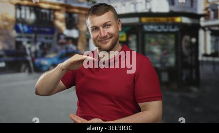 Man in red polo holds hands level as if framing an invisible box on street near kiosk and parked car; confidence ease. Stock Photo