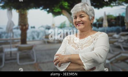 Woman with arms crossed smiling, white lace top and bracelet on a seaside street with loungers and umbrellas; contentment relaxation. Stock Photo