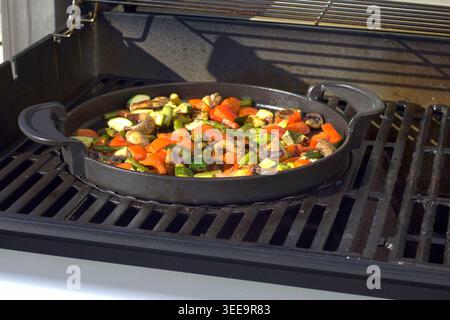 A detailed view of a vegetable stir-fry consisting of mushrooms, red peppers, and zucchini cooking in a seasoned cast iron pan on a barbecue. Stock Photo